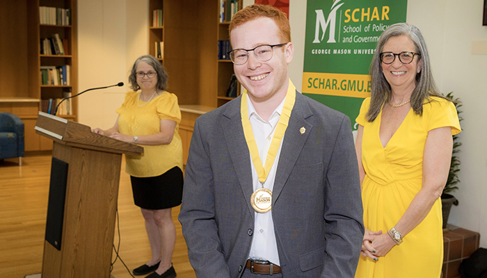 Two women in yellow watch as a man in a gray suit and wearing a gold medal holds a green folder.