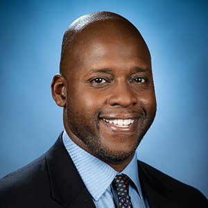 William N. Flagler Jr. stands smiling while wearing a black suit, blue shirt, and tie.