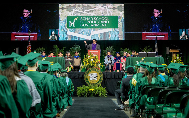 A man in a mortarboard and robe stands at a lectern in front of people wearing green regalia.