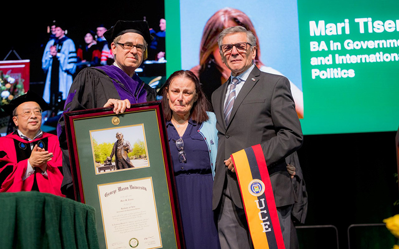 A man in a mortarboard and black robe holds a framed diploma while a woman and man show emotion.