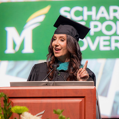A woman with long dark hair and wearing a black mortarboard and robe stands at a podium.